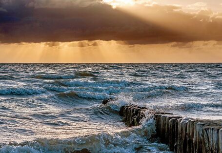 Das Bild zeigt den Ausschnitt eines unruhigen Meeres mit Wellen, die an aus dem Wasser herausragenden Pfählen brechen. Der Himmel ist orange gefärbt und von dunklen Wolken verhangen. Stellenweise treten Sonnenstrahlen zwischen den Wolken hervor.