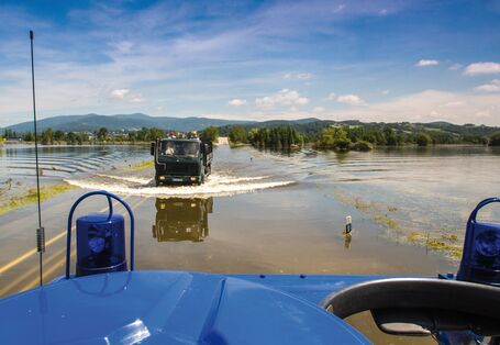 Das Bild zeigt eine von Hochwasser überschwemmte Landschaft. Im Bildzentrum ist eine überschwemmte Straße zu sehen, auf der ein Lastwagen mit Personen auf der Ladefläche fährt. Im Vordergrund des Bildes ist ein Teil eines blauen Einsatzfahrzeugs inklusive blauer Rundumleuchten zu sehen, das ebenfalls auf der überschwemmten Straße unterwegs ist.