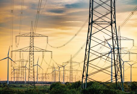 The picture shows numerous high-voltage pylons and power lines against a cloudy, yellowish sky. A few modern wind turbines are visible in the background. 