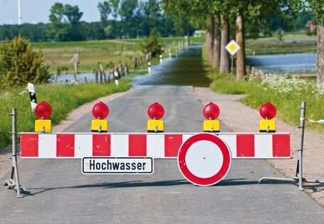 Das Bild zeigt eine durch eine Senke führende überflutete Straße. Im Vordergrund ist sie mit einer Straßensperre abgesperrt. An der Sperre hängt ein Schild mit der Aufschrift "Hochwasser". Rechts und links der Straße sind überflutete Wiesen zu erkennen.