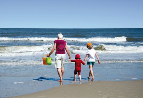 Das Bild zeigt eine ältere Frau, ein Kleinkind und ein Schulkind in Sommerkleidung von hinten. Die drei halten sich an den Händen und laufen am Strand gemeinsam auf die Wellen zu. Die Frau hält zwei Buddel-Eimer in der Hand.