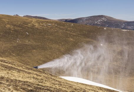 The picture shows an operating snow cannon in a mountainous slope area where there is no snow.