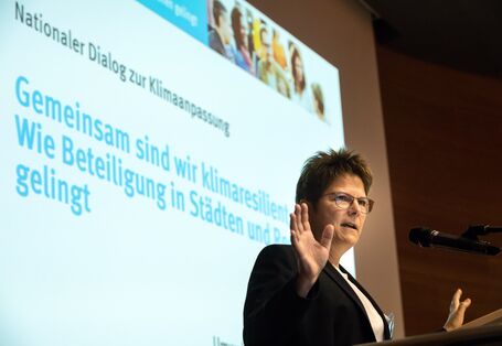 Teilnehmerin steht am Rednerpult, trägt vor und gestikuliert. Im Hintergrund ist der Titel der Veranstaltung zu sehen: Gemeinsam sind wir klimaresilient - Wie Beteiligung in Städten und Regionen gelingt