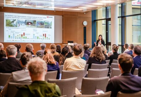 Teilnehmende folgen auf beigen Stühlen in Reihen sitzend einer Präsentation. Die Vortragende steht mit Handmikrofon nahe Stehpult vor einer Fensterfront.