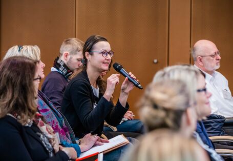 Eine Teilnehmerin spricht angeregt ins Handmikrofon. Alle Teilnehmende sitzen in Reihen im Veranstaltungsraum. Die Wände sind holzvertäfelt.