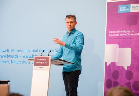 Vortragender in hellblauem Hemd steht gestikulierend am Stehpult vor dem blauen Hintergrund, auf dem der Veranstaltungsort geschrieben steht. Daneben steht das lilafarbene Veranstaltungs-Rollup.