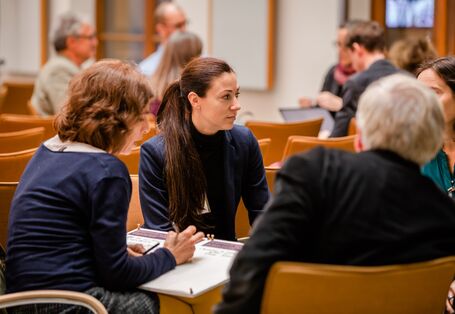 Teilnehmende haben sich in Gruppen im Veranstaltungsraum zusammengesetzt auf orangenen Stühlen und sprechen miteinander. Ergebnisse werden dabei auf Arbeitsbögen von einer Teilnehmerin skizziert.