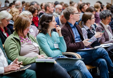Teilnehmende sitzen auf Stühlen in Reihen und beschäftigen sich mit Schriftgut in ihren Händen.