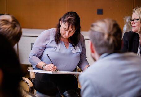 Teilnehmende skizziert auf dem Ergebnisbogen die Ergebnisse aus der Arbeitsgruppe, die im Veranstaltungsraum zusammensitzt. Zwei Teilnehmende tragen hellviolette Oberteile. Der Raum ist holzfarben vertäfelt.
