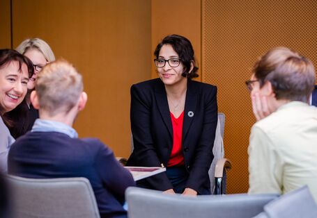 Teilnehmende sitzen in aufgelockerter Stimmung beieinander im holzfarben vertäfelten Veranstaltungsraum. Die Teilnehmerin im Mittelpunkt trägt ein dunkelblaues Sacko mit rotes Oberteil.