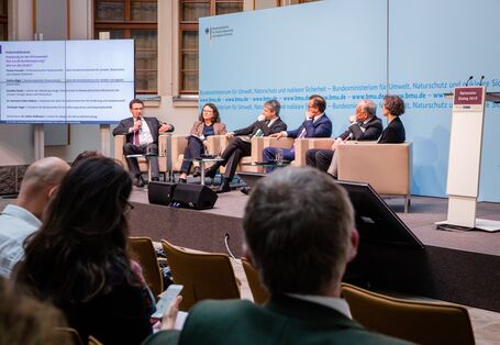 Podiumsdiskussion aus einer anderen Perspektive. Im Hintergrund steht eine Tafel, auf der die Podiumsbeteiligten verzeichnet sind.