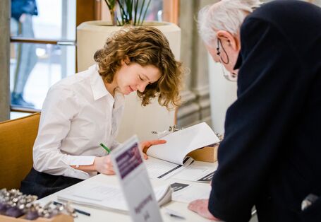 Ein Teilnehmender registriert sich am Registriertisch und trägt sich in die Teilnehmendenliste ein. Eine Dame in einer weißen Bluse trägt Angaben in eine Liste ein.