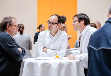 Teilnehmende stehen an weißen Stehtischen in der Pause unterhaltend. Zwei Teilnehmde tragen weiße Hemden. Der helle Raum ist mit gelber Farbe abgesetzt.