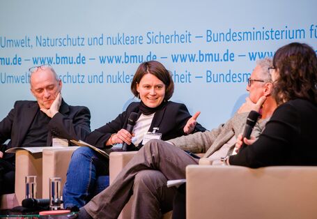 Podiumsdiskussion vor dem hellblauem Hintergrund, auf dem der Veranstaltungsort geschrieben steht. Eine Teilnehmende hält ein Handmikrofon und spricht angeregt zu den anderen Podiumsteilnehmenden.