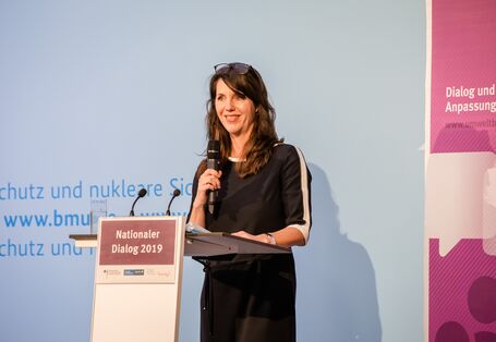 Eine Teilnehmende steht mit Handmikro am Stehpult vor blauem Hintergrund, auf dem der Veranstaltungsort geschrieben steht. Daneben steht das lilafarbene Veranstaltungs-Rollup. Auf dem Pult liegen Unterlagen. Die Teilnehmende schaut lächelnd ins Publikum.