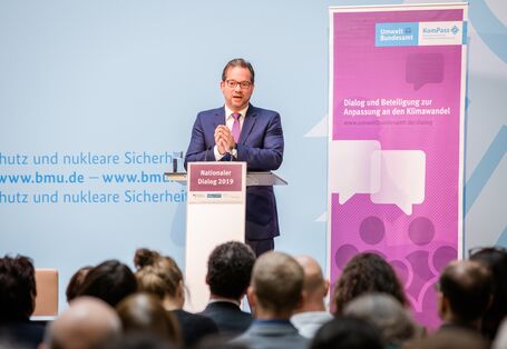 Redner steht am Stehpult und trägt vor. Aufsteller im Hintergrund sowie vorne auf dem Stehpult tragen vorwiegend in lila gehalten den Veranstaltungstitel