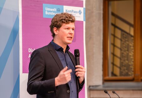 Vortragender steht mit Handmikrofon am Stehpult vor dem lilafarbenen Veranstaltungs-Rollup. Im Hintergrund geht der Blick durch ein hölzernes Fensterkreuz in den Flur des Gebäudes.