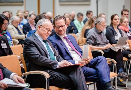 Teilnehmende sitzen auf Stühlen in Reihen und beschäftigen sich mit Schriftgut in ihren Händen.