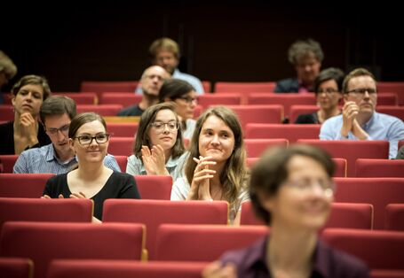 Teilnehmerinnen und Teilnehmer sitzen applaudierend in den roten Stuhlreihen des Hörsaales.