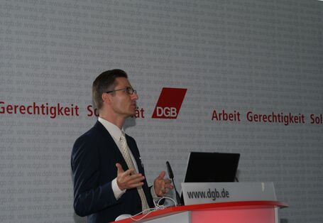Vortragender steht gestikulierend am Rednerpult. Im Hintergrund ist das Logo vom DGB Arbeit Gerechtigkeit Solidarität zu sehen.