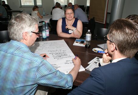 Teilnehmer sitzen im Arbeitsgruppenraum am Arbeitsgruppentisch und diskutieren miteinander.