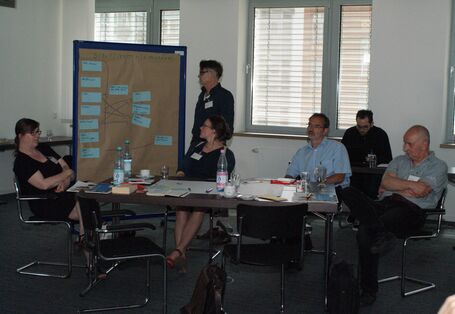 Teilnehmer sitzen nachdenklich am Arbeitsgruppentisch. Im Hintergrund schaut sich ein Teilnehmer die Moderationskarten auf der Moderationswand an.