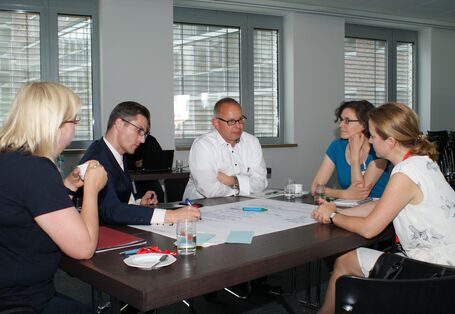 Teilnehmer diskutieren am Gruppenarbeitstisch. Ein Teilnehmer schreibt die Diskussionsergebnisse auf Posterpapier, welches auf dem Tisch liegt.