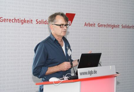 Vortragender steht am Rednerpult vor aufgeschlagenem Laptop. Im Hintergrund ist das DGB Logo zu sehen: Gerechtigkeit, Solidarität, Arbeit