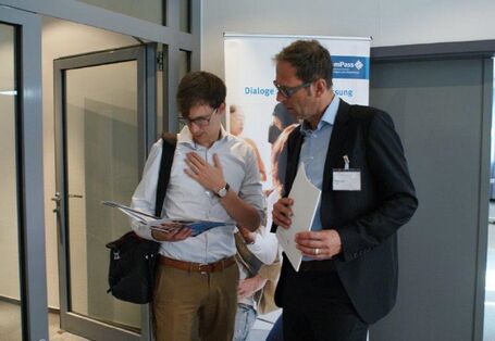 Zwei Teilnehmer stehen zusammen und schauen vertieft in die Veranstaltungsbroschüre. Im Hintergrund ist das Veranstaltungs Rollup sowie eine geöffnete Glastür zum Flur hin zu sehen.