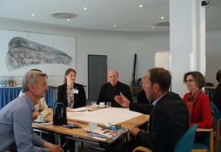 Teilnehmer sitzen diskutieren am Arbeitsgruppentisch und hören dem gestikulierenden Sprecher zu. Alle schauen freundlich drein.