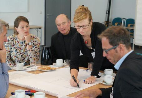 An einem Arbeitsgruppentisch steht eine Teilnehmerin und schreiben mit einem schwarzen Stift einen Beitrag auf Papier. Die anderen Teilnehmer schauen interessiertzu.