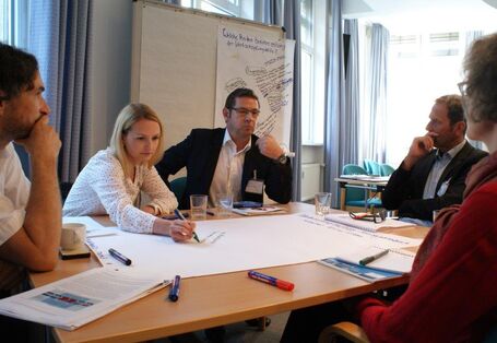 Teilnehmer sitzen an einem Gruppenarbeitstisch. Eine Teilnehmerin schreibt die ausgesprochenen Gedanken eines anderen Teilnehmers auf Papier. Andere Teilnehmer folgen dem interessiert.