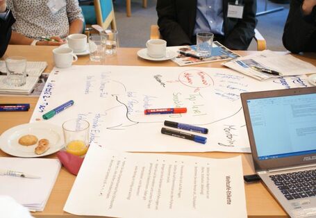 Ansicht auf einen Gruppenarbeitstisch zeigt in bunten Farben beschriebenes Papier, Stifte, ein aufgeklapptes Laptop, einen Teller mit Keksen und Gläser und Tassen mit Getränken.