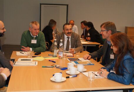 Teilnehmende sitzen am Gruppenarbeitstisch und sprechen miteinander. Auf dem Tisch liegen Moderationsmaterialien wie Stifte und farbige Moderationskarten sowie Getränke. Im Hintergrund sieht man andere Teilnehmende ebenfalls am Gruppenarbeitstisch diskutierend. 