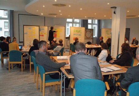 Im Vordergrund sitzen die Teilnehmenden auf verschiedene Arbeitsgruppentischen verteilt im Raum und hören vertieft dem Vortragenden zu. Dieser steht vor einigen Moderationswänden, an denen beschriftete Moderationskarten hängen und zeigt erläuternd auf die Moderationswand. Der Blick geht im Hintergrund durch Fenster auf eine Häuserwand. 