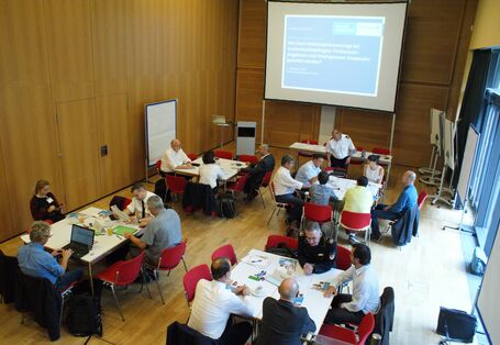 Die Aufnahme zeigt von oben die Teilnehmenden an Gruppenarbeitstischen sitzend in einem mit Holz vertäfelten Raum. Im Hintergrund ist das Veranstaltungsmotto Katastrophenschutz bei Trockenheit in der Landwirtschaft  in blau/weiss auf der Leinwand projiziert zu sehen.