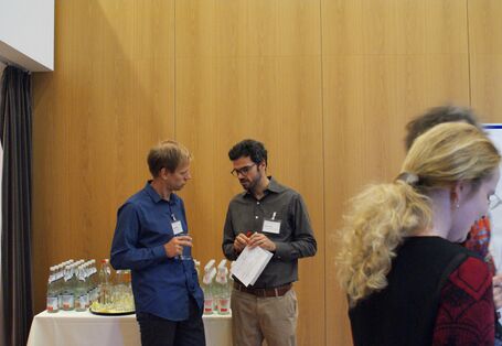 Zwei Teilnehmende stehen vor dem Getränketisch und unterhalten sich. Beide tragen Namenschilder am Hemd und halten Papiere und ein Glas Wasser in den Händen.