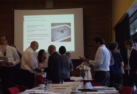 Die Teilnehmenden stehen im für Präsentationszwecke verdunkelten Veranstaltungsraum in lockeren Gruppen beieinander. Im Hintergrund ist eine Präsentation zu sehen.