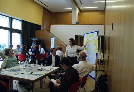 Im Vordergrund steht Gruppenarbeitstisch 3. Die Teilnehmenden sitzen um den Tisch herum und folgen den Ausführungen einer Teilnehmerin, die an der Präsentationswand zeigend steht. Im Hintergrund befindet sich eine große Fensterfront.