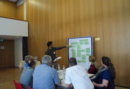 Ein Teilnehmer zeigt auf eine Präsentationswand mit grünen Moderationskarten. Im Vordergrund sitzen Teilnehmende am Gruppenarbeitstisch und hören zu.