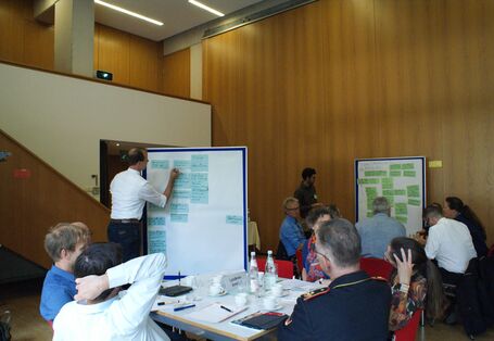 Ein Teilnehmender schreibt etwas auf eine Präsentationtswand, die mit blauen Moderationskarten bestückt ist. Im Raum sitzen alle Teilnehmenden an ihren Gruppenarbeitstischen.