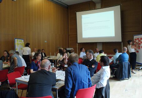 Der Veranstaltungsraum im Überblick zeigt alle Teilnehmenden an ihren Gruppenarbeitstischen und schauen zu, wie eine Teilnehmerin an einer Präsentationswand mit roten Moderationskarten präsentiert und etwas anschreibt. Auf der gegenüber liegenden Seite steht eine Präsentationswand mit gelben Moderationskarten.