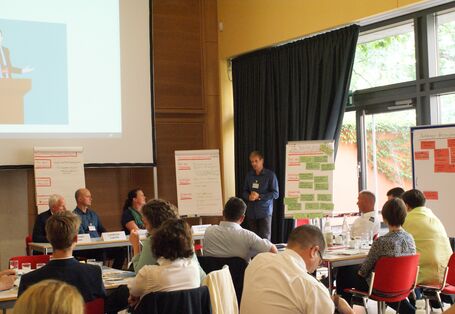 Die Teilnehmenden sitzen an ihren Gruppenarbeitstischen vor den aufgereihten Präsentationswänden,die mit den Arbeitsergebnissen anhand von farbigen Moderationskarten und Schriften bestückt sind. Das Licht im Raum wird von der gelben Wand an der Fensterfront reflektiert und erscheint sehr hell und freundlich.