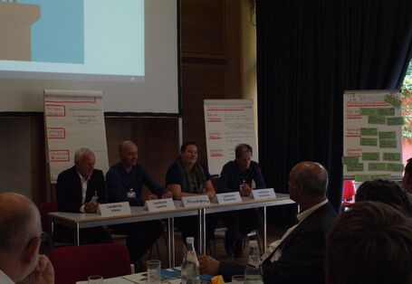 Auf einem Podium sitzen die teilnehmenden Vertreter der  Landkreise aufgereiht an einer Tischreihe. Namensschilder und Getränke stehen auf dem Tisch. Im Hintergrund stehen verschiedene Präsentationswände. Der Veranstaltungsraum ist präsentationsbedingt verdunkelt.