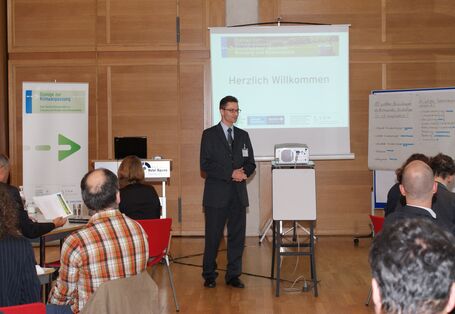 Vortragender begrüßt die Teilnehmer. Seine Hände sind gefalten. Im Hintergrund stehen Moderationswände, der Veranstaltungsaufsteller Dialoge zur Klimaanpassung und eine Leinwand mit dem Schriftzug Herzlich Willkommen.