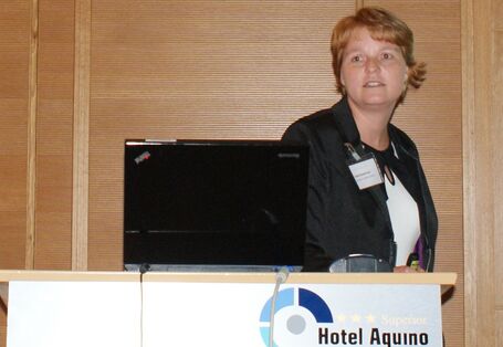 Vortragende steht vor holzvertäfelten Wand am Rednerpult. Das Rednerpult trägt vorne den Schriftzug Hotel Aquino Tagungszentrum.