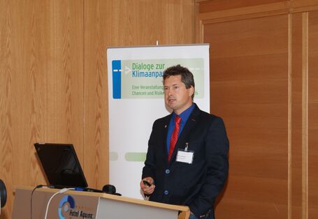 Vortragener steht am Rednerpult. Ein Laptop mit geöffnetem Deckel steht auf dem Rednerpult. Im Hintergrund steht der Veranstaltungsaufsteller Dialoge zur Klimaanpassung.