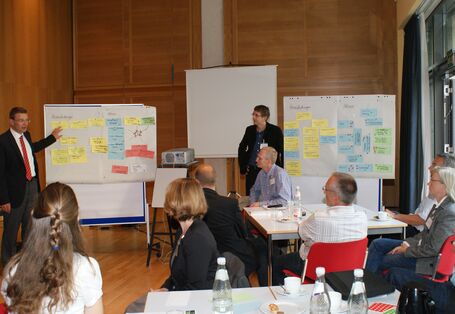 Teilnehmer sitzen an Gruppenarbeitstischen und hören Vortragenden zu. Vortragende stehen im Hintergrund vor Moderationswänden, die mit bunten Moderationskarten bestückt sind. Von der rechten Seite her scheint Tageslicht durch die Fensterfront.