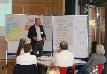 Vortragender steht vor Moderationswänden und präsentiert Ergebnisse. Im Vordergrund sitzen die Zuhörer am Gruppenarbeitstisch.