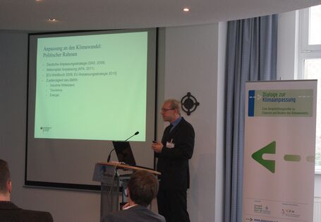 Vortragender steht am Rednerpult. Im Hintergrund ist die Präsentation auf Projektionswand zu sehen und der Veranstaltungsaufsteller.